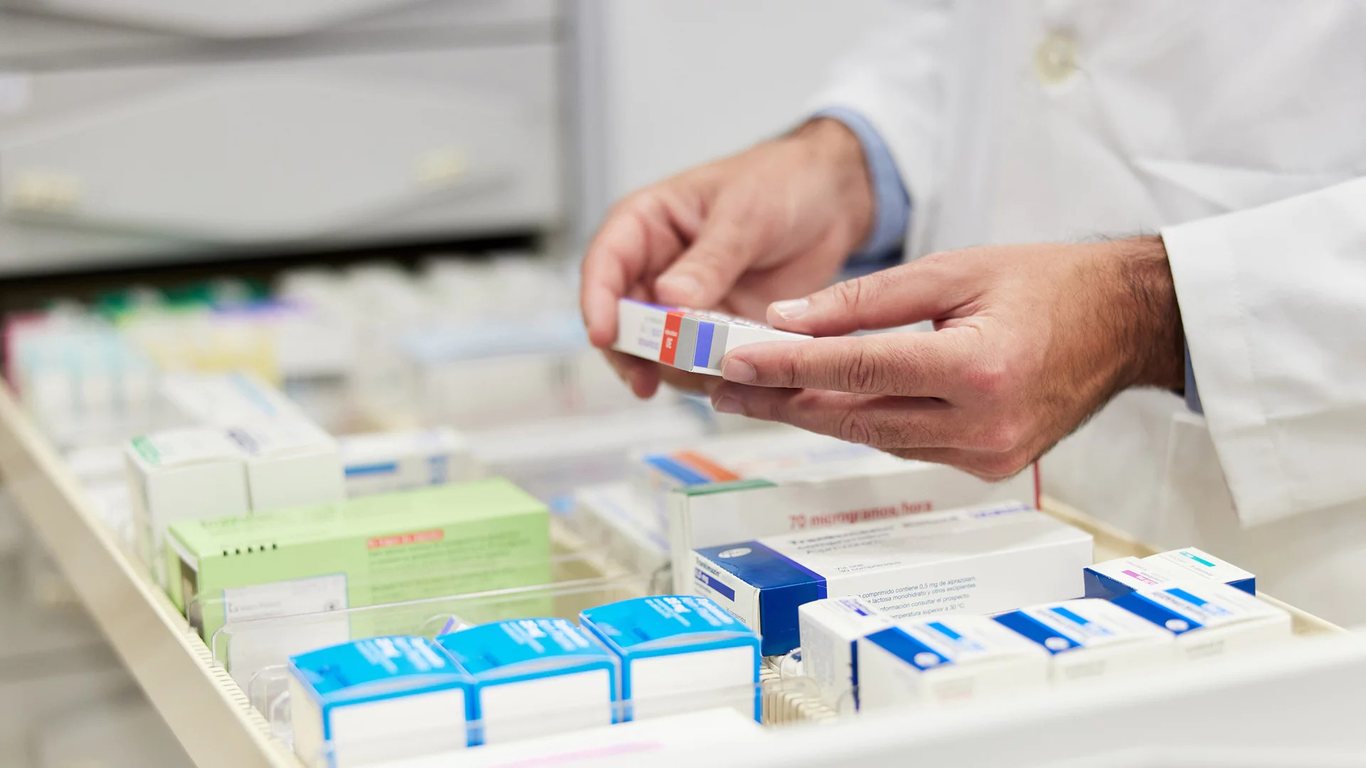 Fotografie zeigt Hände in weißem Kittel, die eine Medikamentenverpackung aus einem Schubladensystem mit verschiedenen Arzneimitteln entnehmen. Fokus liegt auf der Organisation und Auswahl von Medikamenten in einer Apotheke oder Klinik.