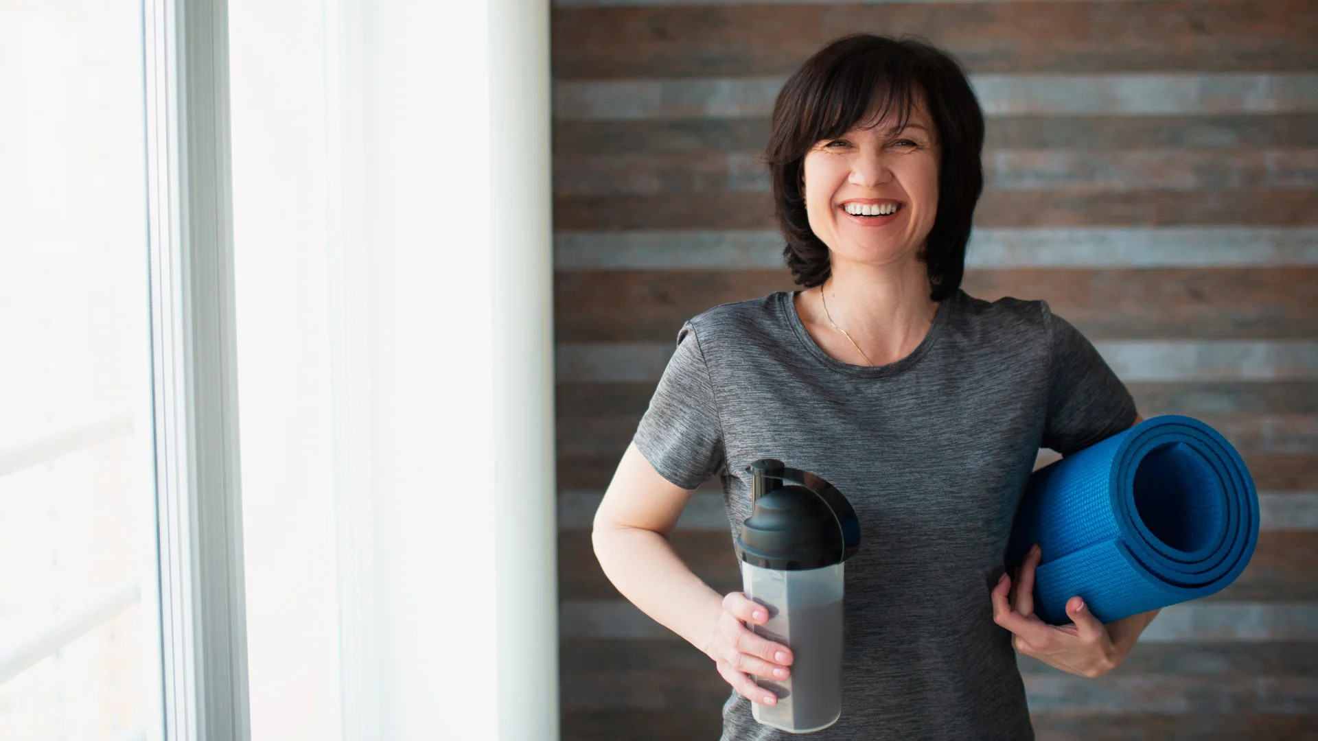 Fotografie einer lächelnden Frau in Sportkleidung, die eine blaue Yogamatte unter dem Arm hält und einen Proteinshake in der Hand hat. Szene vermittelt Vorbereitung auf Fitness- oder Yoga-Training in hellem Raum mit Holzwand im Hintergrund.