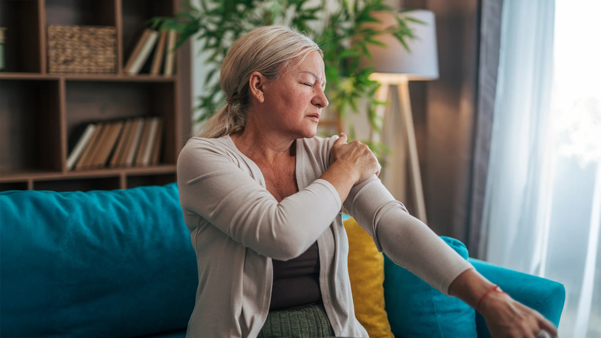 Fotografie einer älteren Frau, die auf einem Sofa sitzt und sich wegen Schulterschmerzen an den Oberarm greift.