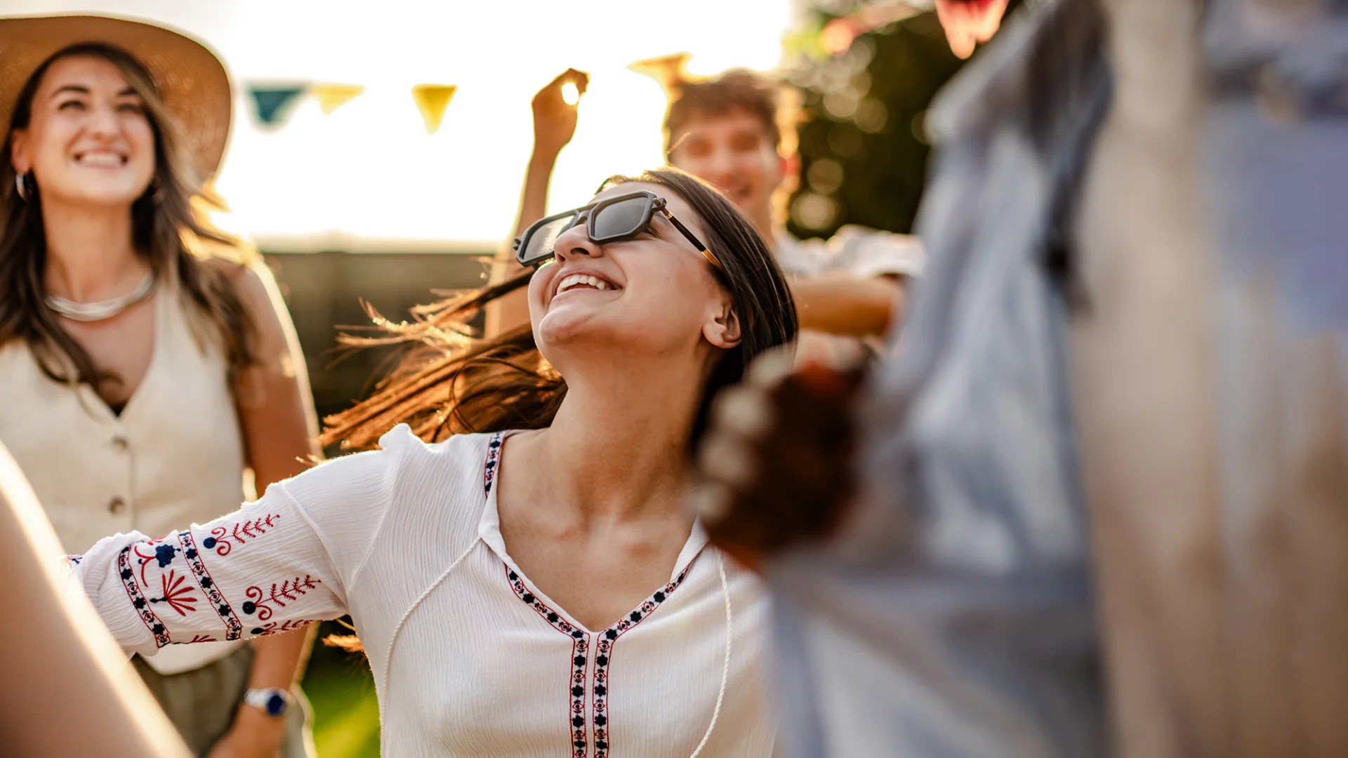Fotografie von mehreren fröhlichen Menschen bei sonnigem Wetter, die gemeinsam im Freien feiern. Im Vordergrund eine junge Frau mit Sonnenbrille und weißer Bluse, die lächelnd nach oben blickt, umgeben von weiteren lächelnden Personen.