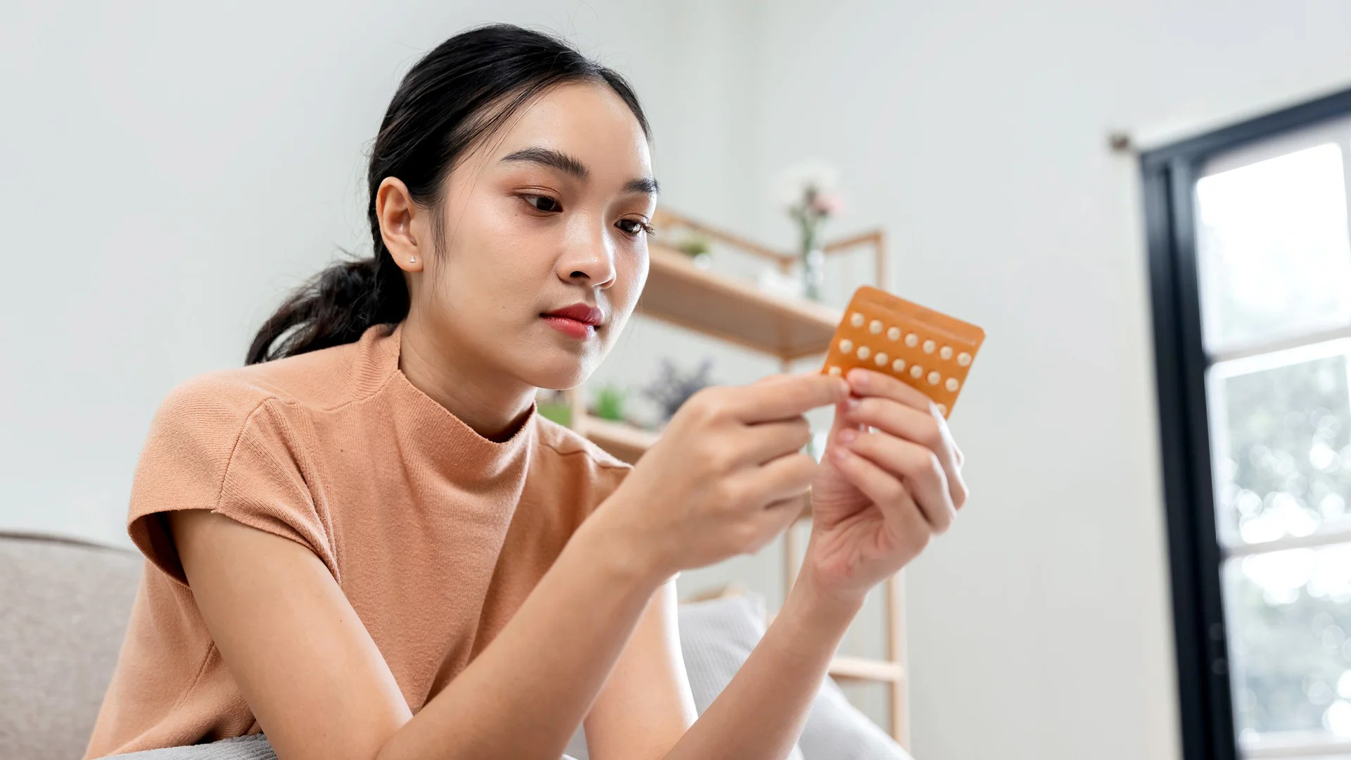 Fotografie einer jungen Frau, die aufmerksam eine orangefarbene Medikamentenpackung mit Tabletten betrachtet.